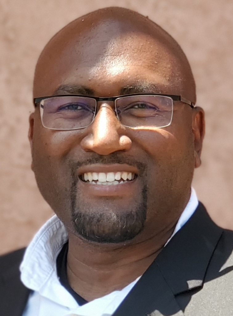 Close up image of bald man with closely cropped facial hair and glasses wearing a suit jacket smiling