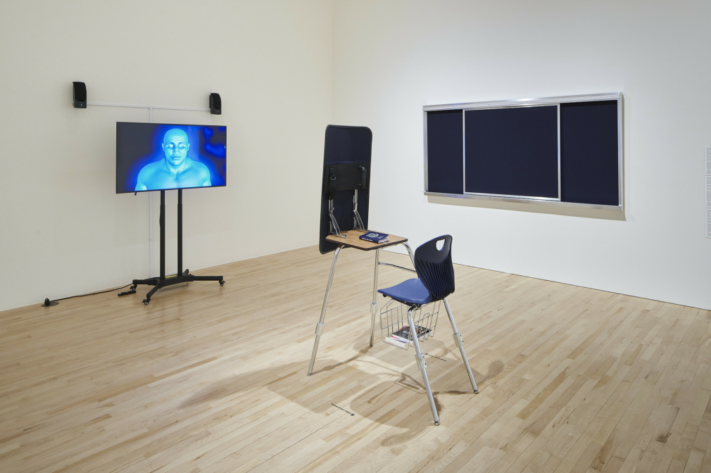 A single school desk and chair face a flat screen TV with an image of a blue man on it.