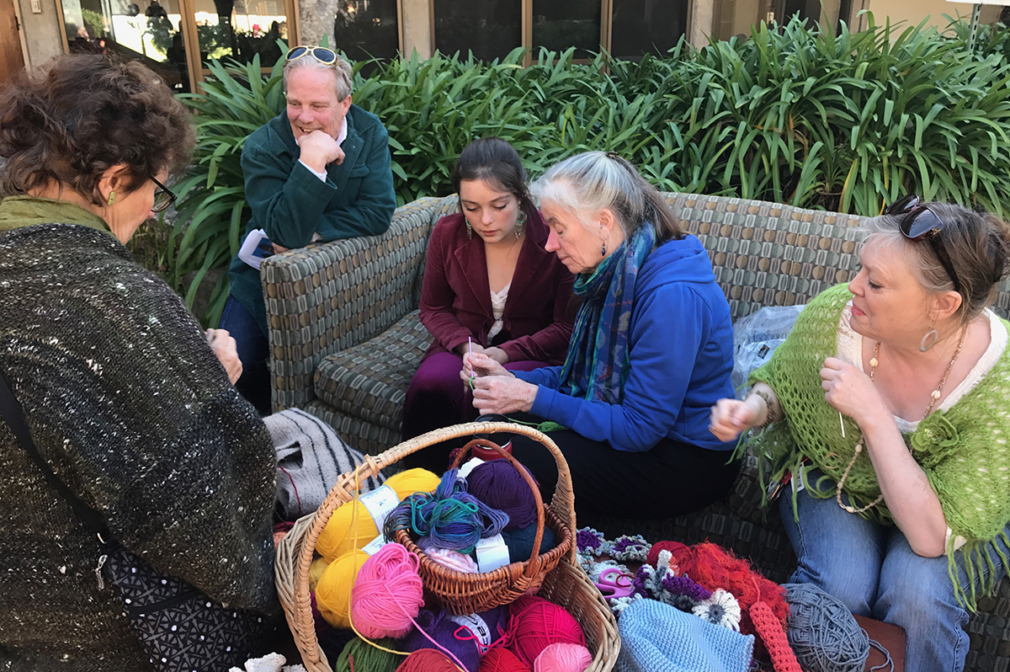 A group of people sit on an outdoor couch, crocheting.
