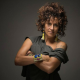 A woman with curly brown hair and a gray shirt poses with her arms crossed.