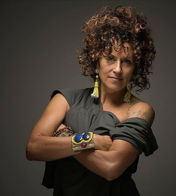 A woman with curly brown hair and a gray shirt poses with her arms crossed.