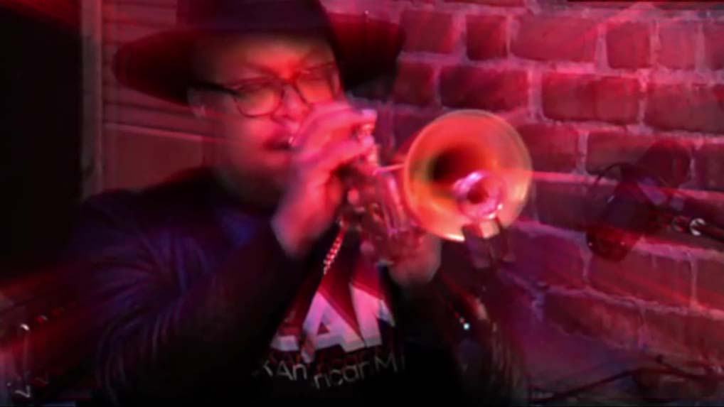 A black man plays a trumpet in front of a brick wall.