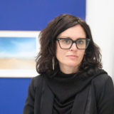 woman with dark hair , heavy black glasses frames, and dressed in all black leans against a white wall.