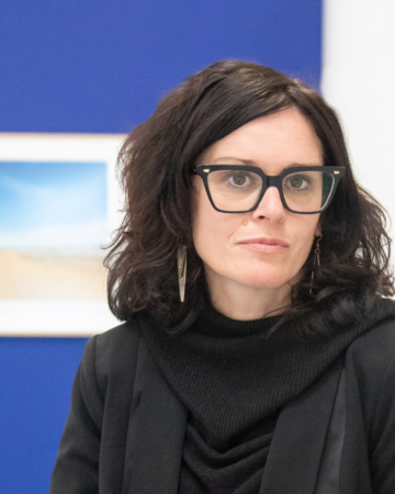 woman with dark hair , heavy black glasses frames, and dressed in all black leans against a white wall.