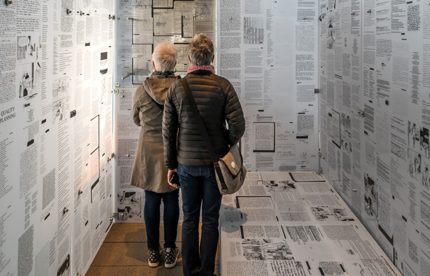 Two women stand in an exhibit consisting of writings plastered to the walls and floor.