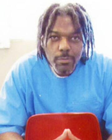 A black man sits in a red chair in a prison cell.