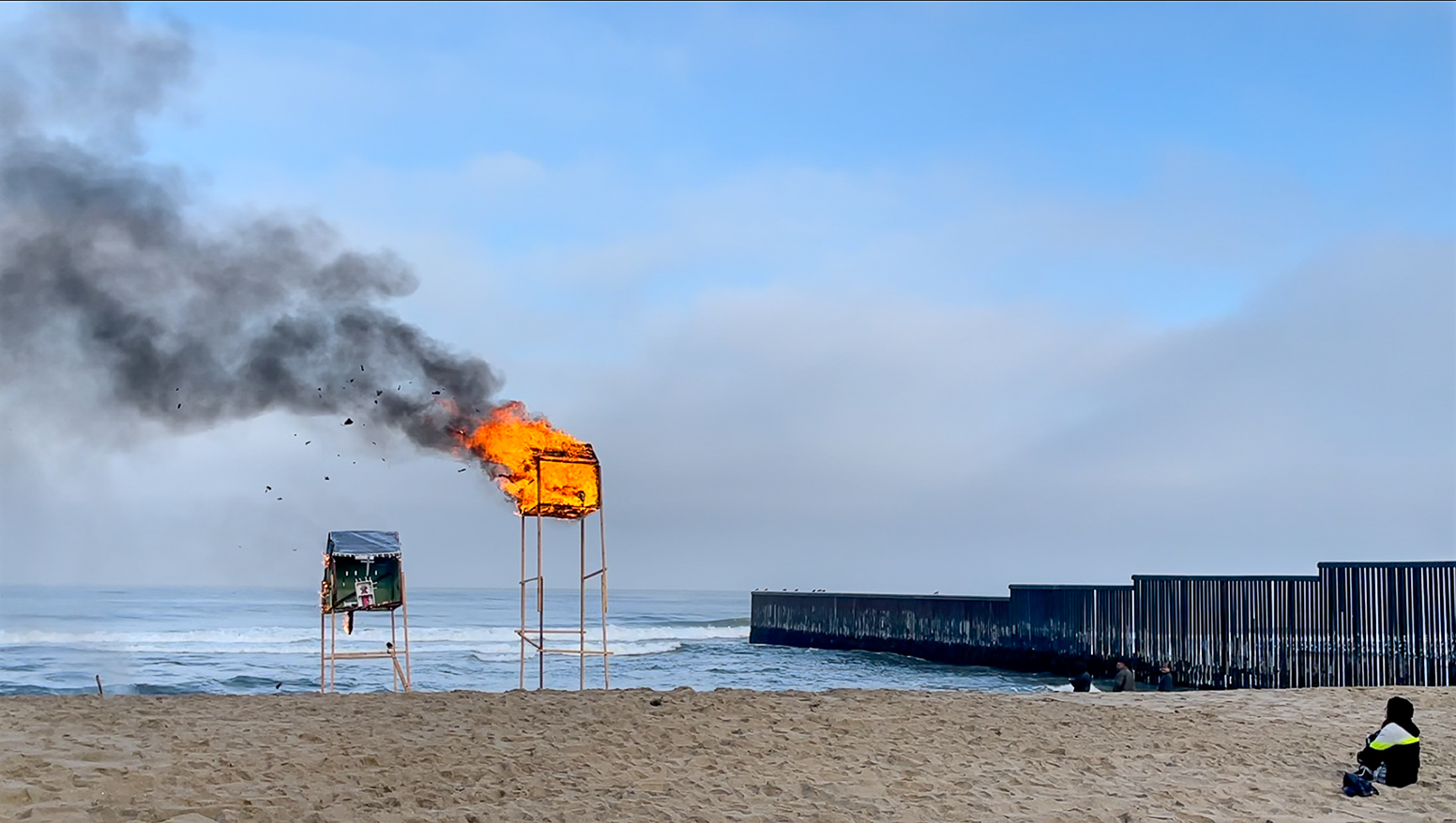 A person sits on a beach watching as a lifeguard stand burns.