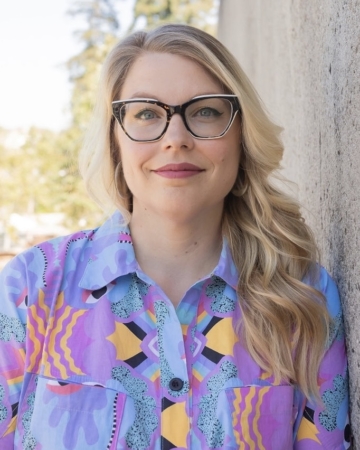Person with long blond hair, glasses, and blue eyes wearing a bright, patterned shirt