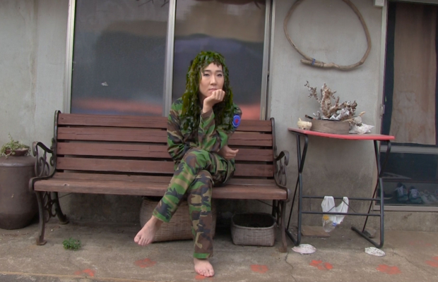 A woman in military fatigues and bare feet sits on a bench and faces the camera.