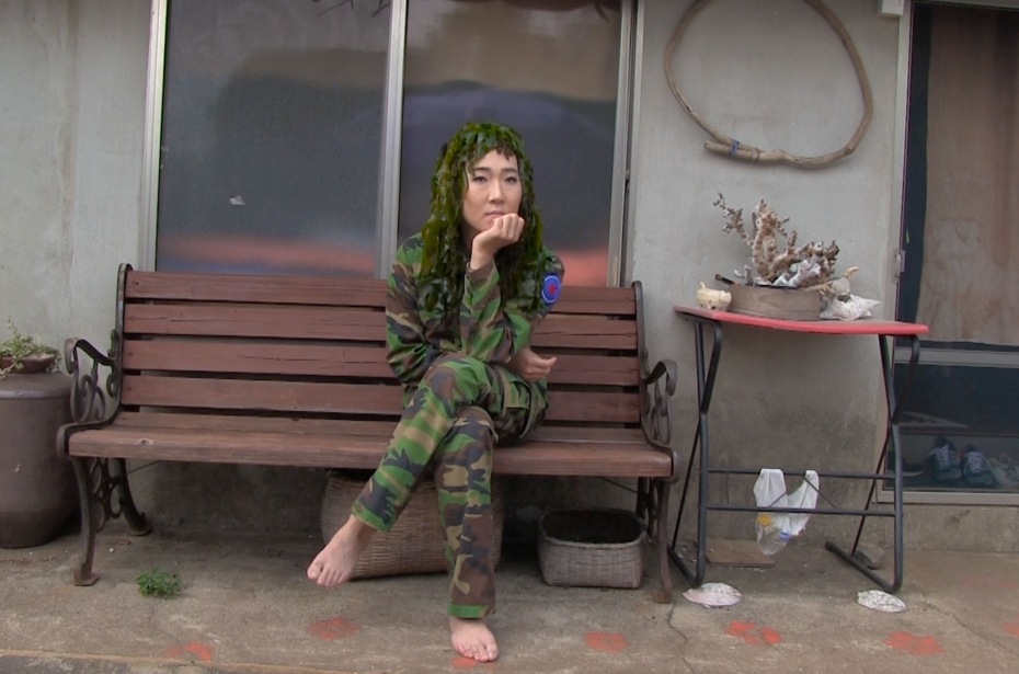 A woman in military fatigues and bare feet sits on a bench and faces the camera.