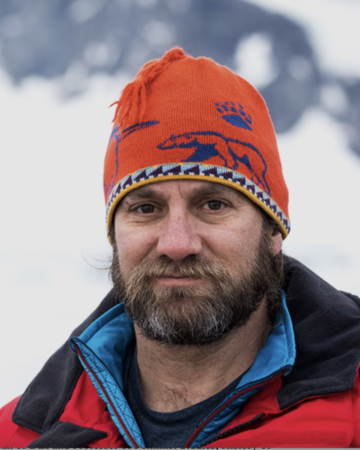 bearded man with orange snow cap and orange jacket in front of iceburg