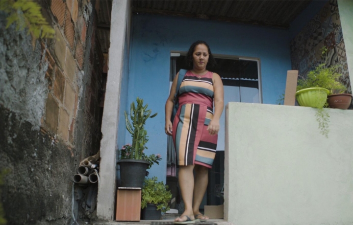 Still from Rioters. A woman in a striped dress, exits a porch.